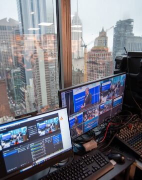 The view through a window in a skyscraper in the NewYork looking across the skyline. In the foreground there is a livestream audio and video mixing desk.