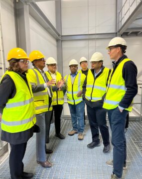 a group of people in yellow high viz vests and hard hats are talking in a factory.