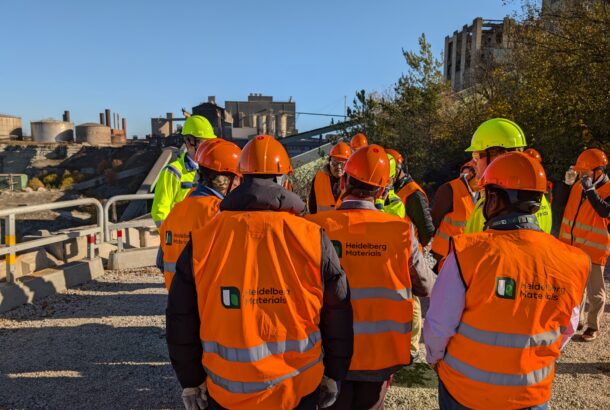 A group in orange high viz vests and hard hats are standing with their backs to camera looking at a cement factort.